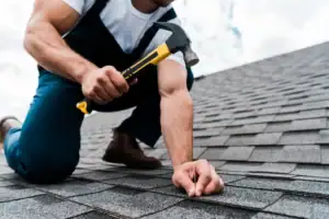 A Roofing Contractor Delaware works on a roof, hammering asphalt shingles into place during installation.